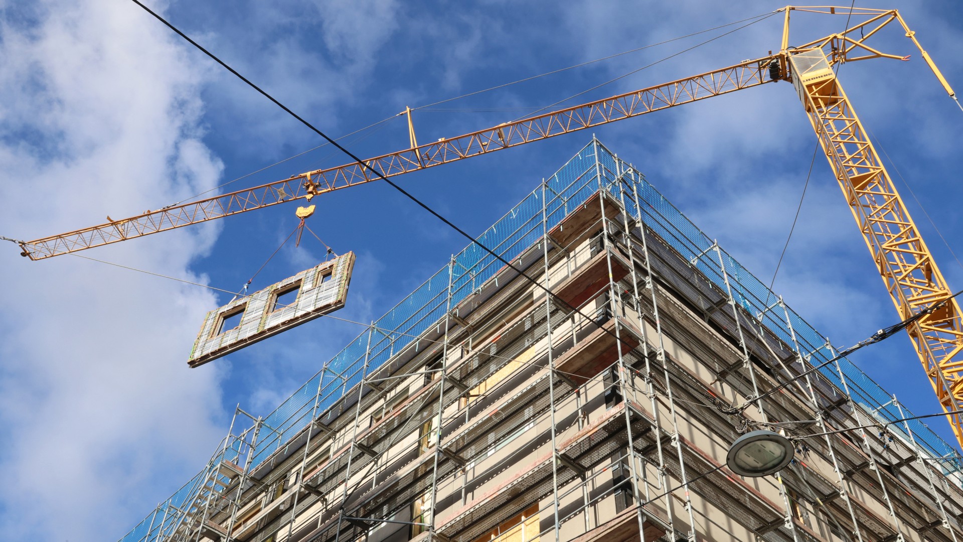 Ein mehrstöckiges Gebäude im Bau mit Gerüst. Ein Kran hebt ein vorgefertigtes Fassadenelement.