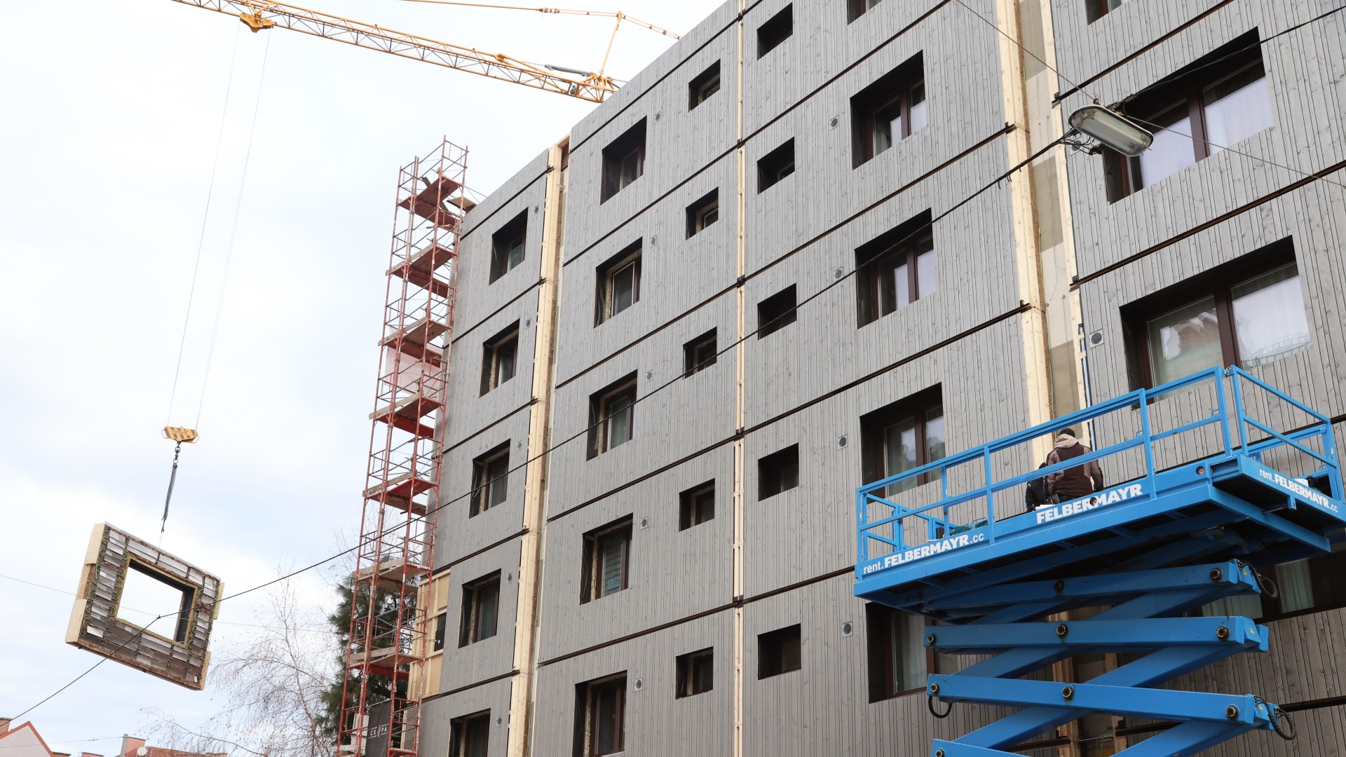 © Klimafonds / Krobath Ein mehrstöckiges Gebäude im Bau mit rotem Gerüst am linken Gebäuderand. Ein Kran hebt ein vorgefertigtes Fassadenelement und zwei Personen stehen auf einem blauen Hubsteiger.