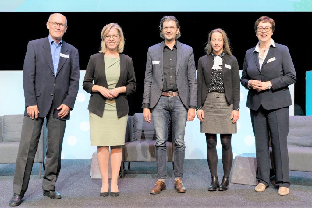 Fünf Personen stehen vor drei Sesseln und einem Teil einer Präsentation, die auf der Wand angezeigt wird.