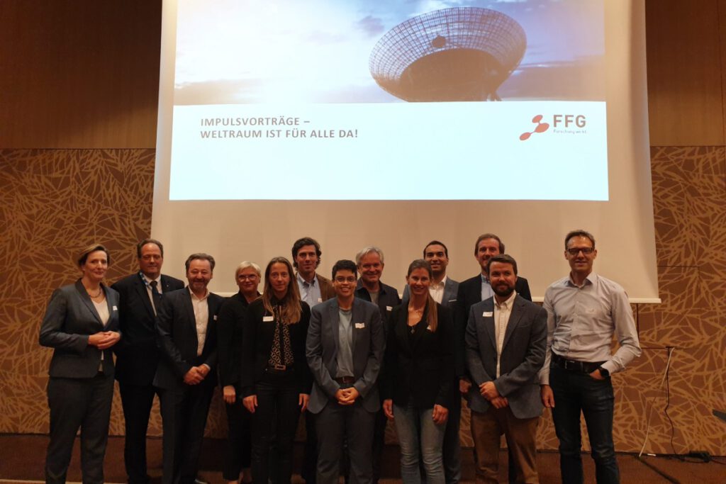 Eine Gruppe von dreizehn Personen steht vor einer weißen Leinwand, auf der mittels Beamer eine Präsentation angezeigt wird