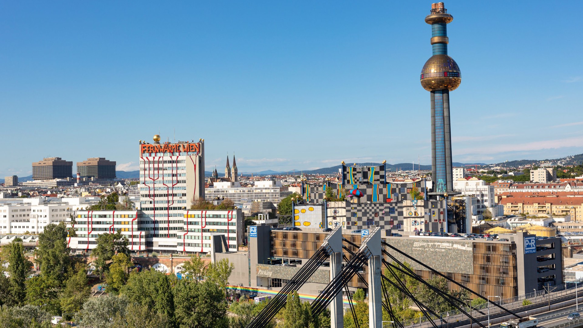 Stadt mit hohem Turm einer Müllverbrennungsanlage mit bunt verzierten Gebäuden rundherum und einem Gebäude der Fernwärme Wien daneben