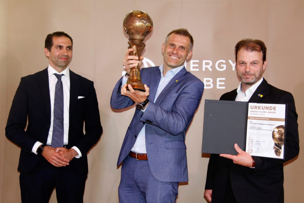 Drei Personen stehen vor einer Wand. Die Person rechts hält eine Urkunde des Energy Globe Award Österreich 2023 in der Hand. Die mittlere Person hält einen Pokal in der Hand. Die Person rechts sieht den Pokal an.