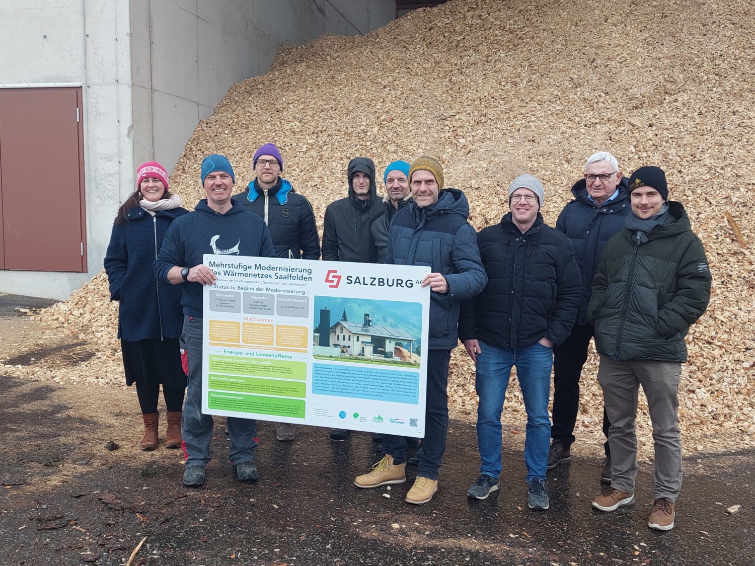 Eine Gruppe von neun Personen steht vor einem Haufen Hackschnitzel und hält eine Schautafel hoch