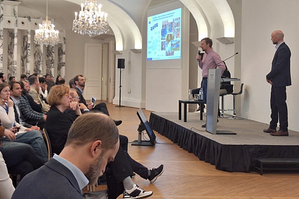 Zwei Männer stehen auf einem Podium und einer von ihnen hält ein Mikrofon in der Hand. Unten sitzt eine Menge von Personen.