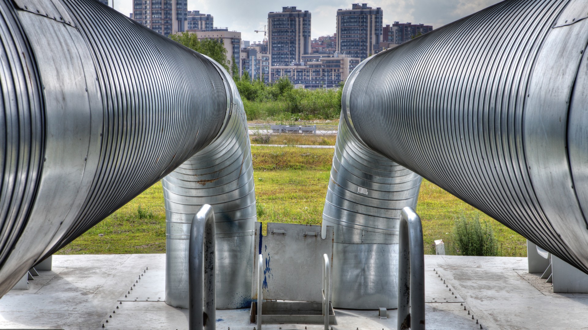 © iStock.com/Grigorev_Vladimir Zwei große, silberne Metallrohre führen in eine Betonplattform mit Treppengeländer mit Hochhäusern im Hintergrund