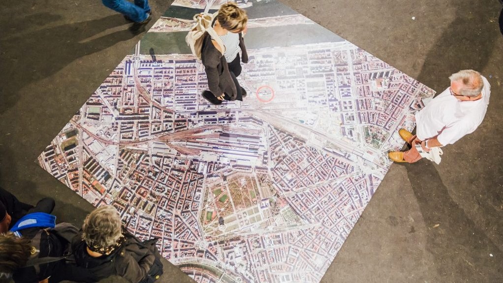 © Wien wird WOW Eine Person steht auf einem auf dem Boden liegenden Bild einer Stadt aus Vogelperspektive und drei weitere Personen stehen am Rand des Bildes, aufgenommen aus der Vogelperspektive