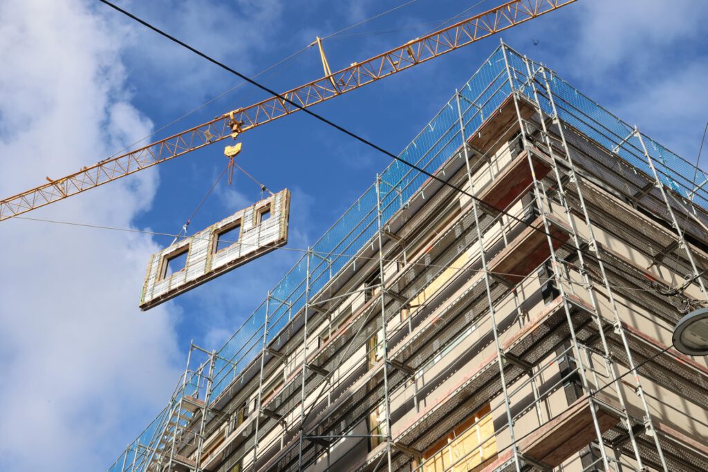 Fassadenelement wird mit Kran zu einem Gebäude transportiert, das von einem Baugerüst umgeben ist