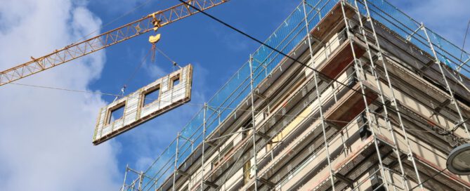 Fassadenelement wird mit Kran zu einem Gebäude transportiert, das mit einem Baugitter umgeben ist