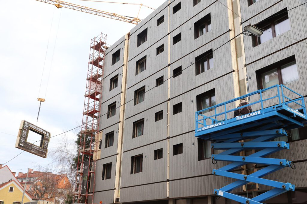 Ein mehrstöckiges Gebäude im Bau mit rotem Gerüst am linken Gebäuderand. Ein Kran hebt ein vorgefertigtes Fassadenelement und zwei Personen stehen auf einem blauen Hubsteiger.