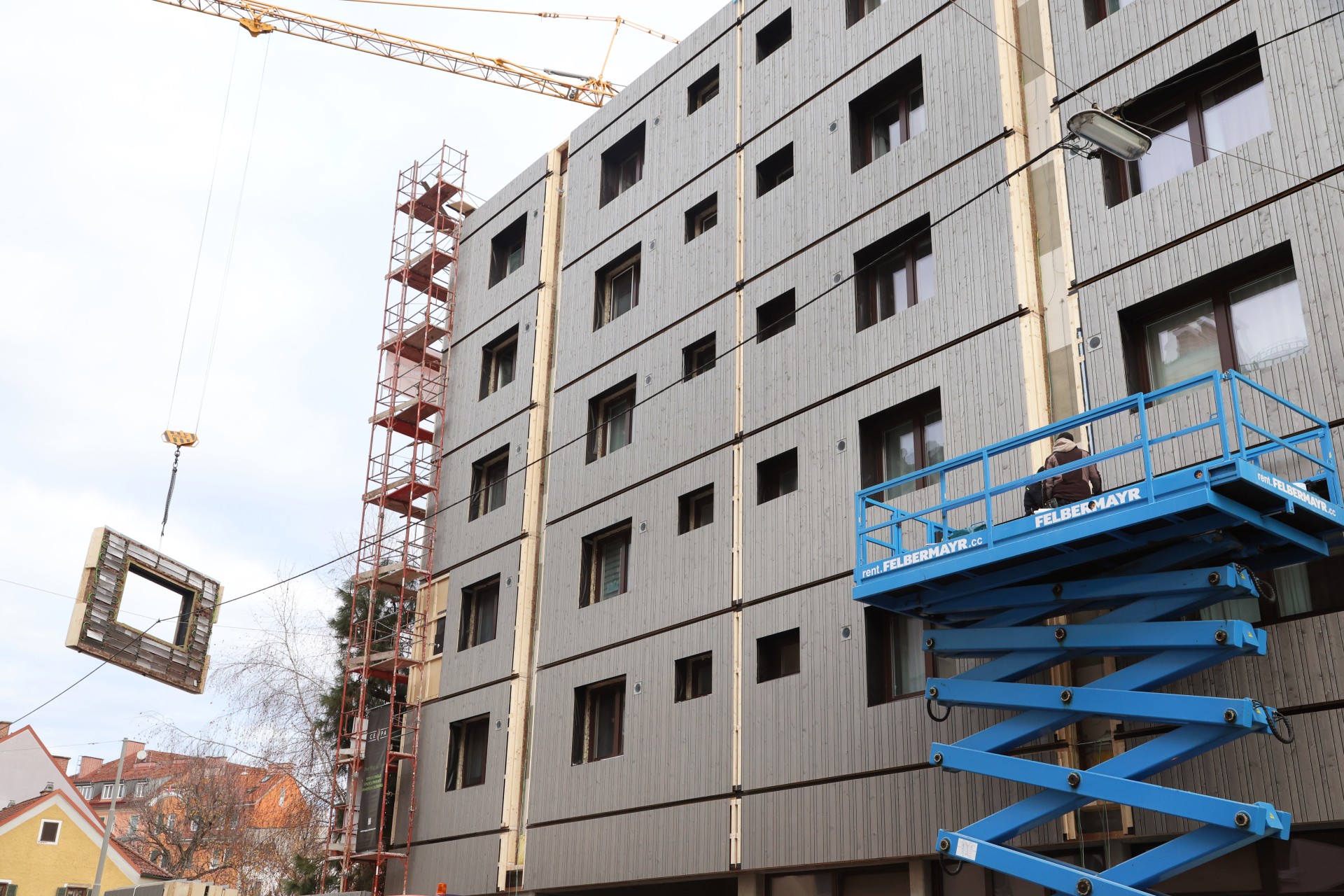 Ein mehrstöckiges Gebäude im Bau mit rotem Gerüst am linken Gebäuderand. Ein Kran hebt ein vorgefertigtes Fassadenelement und zwei Personen stehen auf einem blauen Hubsteiger.