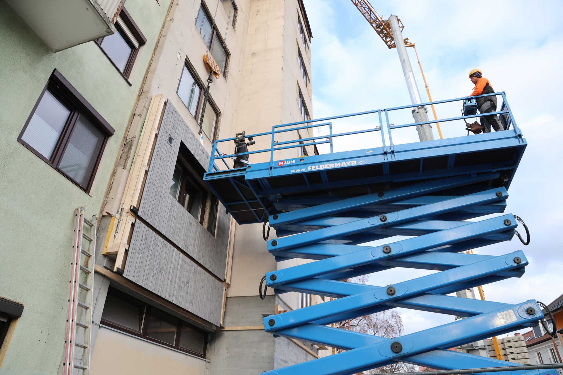 Ein mehrstöckiges Gebäude im Bau. Ein Kran hebt ein vorgefertigtes Fassadenelement an die Wand und zwei Personen stehen auf einem blauen Hubsteiger.