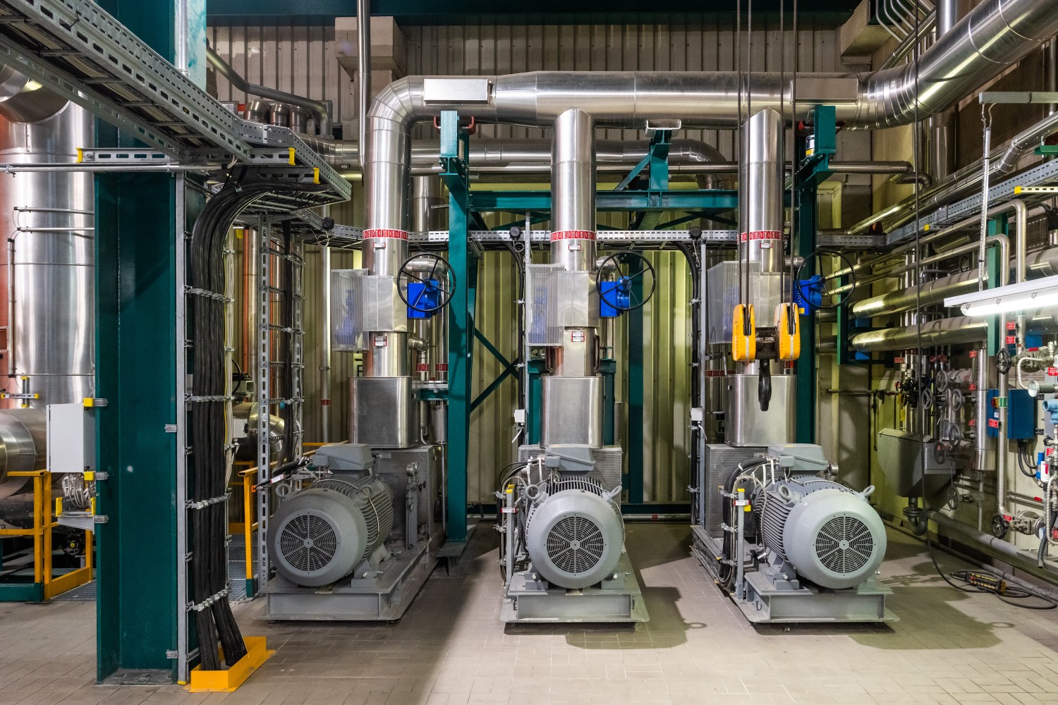 Drei industrielle Maschinen mit Elektromotoren und vertikalen Rohrleitungen in einer technischen Anlagehalle.