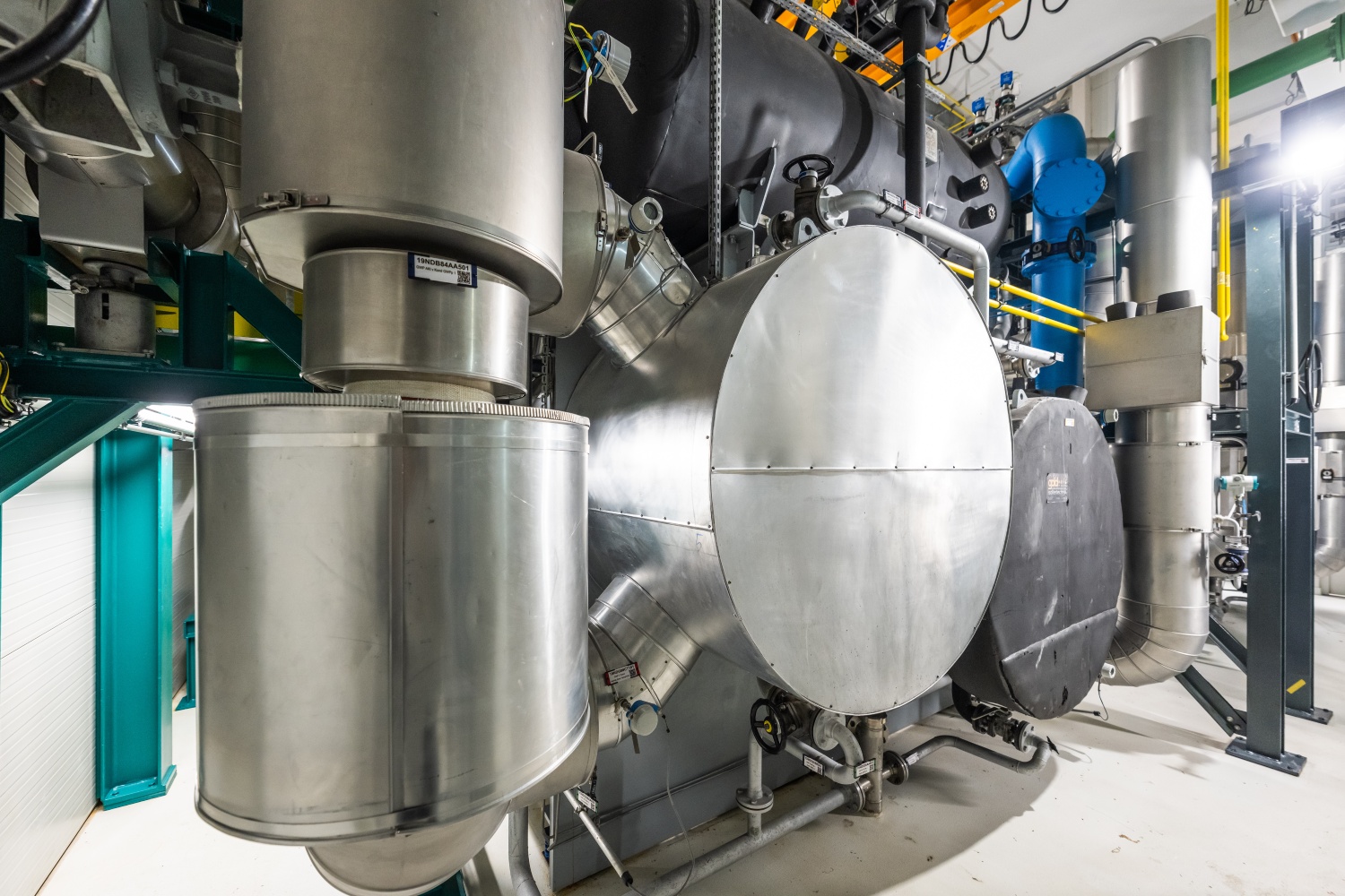 Zwei Großwärmepumpen aus Edelstahl mit angeschlossenen Rohrleitungen und Tanks in einem Technikraum.