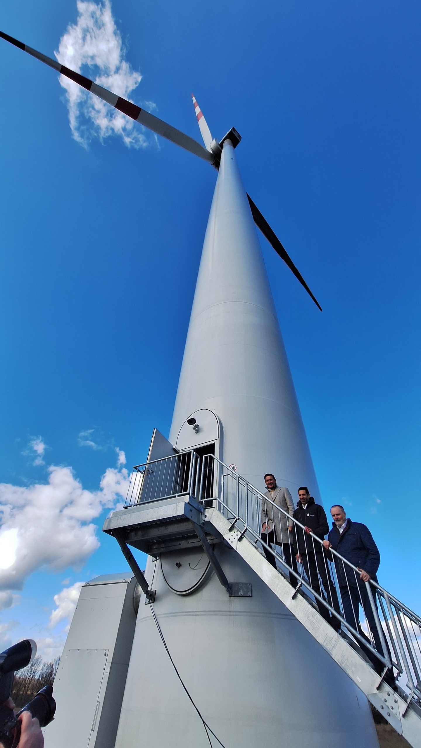 Ein Windrad aus der Froschperspektive fotografiert. Drei Personen stehen auf einer Stiege, die ins Windrad führt.