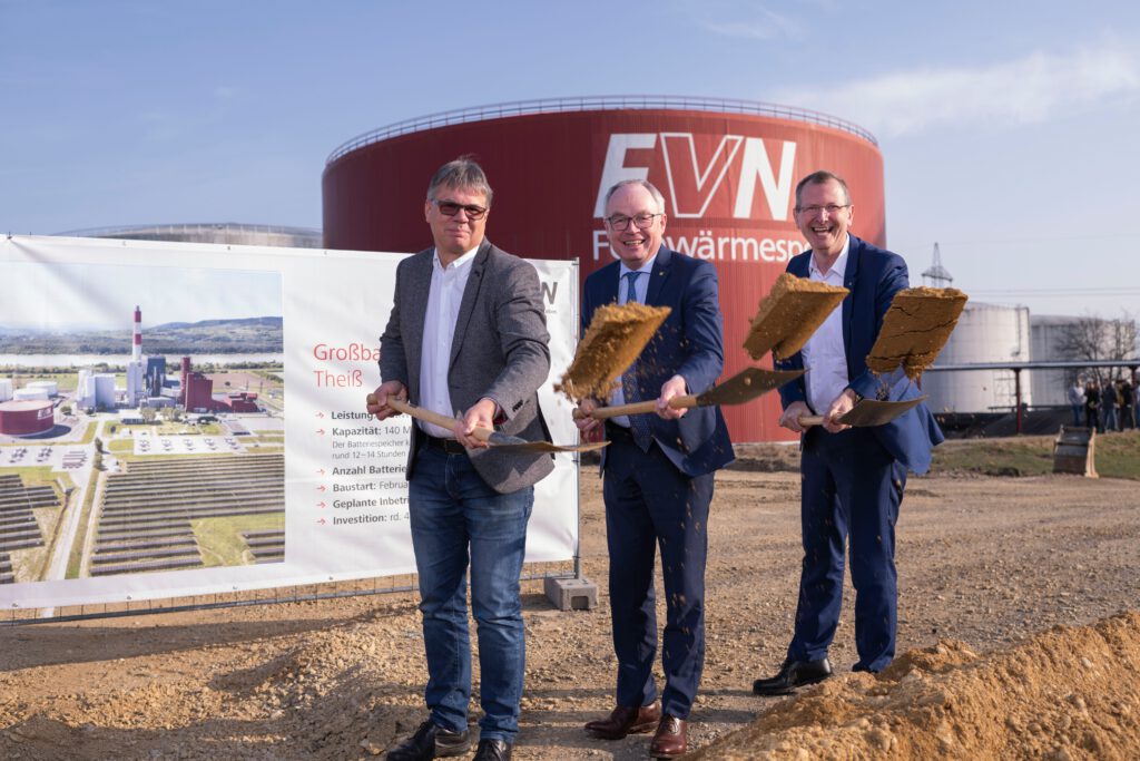 Drei Personen stehen vor dem EVN Fernwärmespeicher und einem Schild mit Informationen zum Großbatteriespeicher Theiß. Die Personen halten jeweils eine Schaufel in der Hand und werfen Erde in die Luft.
