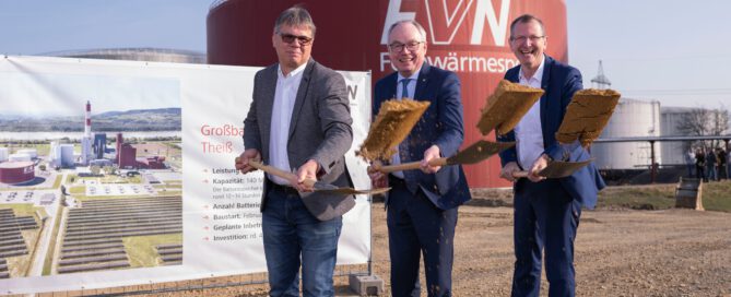 Three people are standing in front of the EVN district heating storage facility and a sign providing information about the Theiss large-scale battery storage facility. Each of them is holding a shovel and throwing earth into the air.