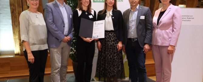 Six people stand side by side on a stage, smiling at the camera. A woman in the centre holds an open award certificate folder, with an ÖGUT banner in the background.